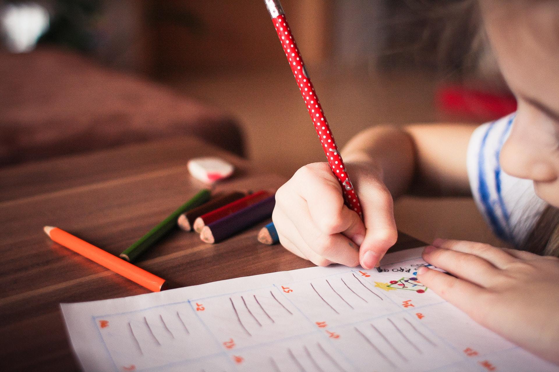 Ein Kind schreibt mit einem roten Bleistift auf einem Blatt Papier, umgeben von bunten Stiften auf einem Holztisch – eine kreative Lernaktivität, die demokratische Werte und Vielfalt fördert, wie VIDETO sie für Kinder unterstützt.