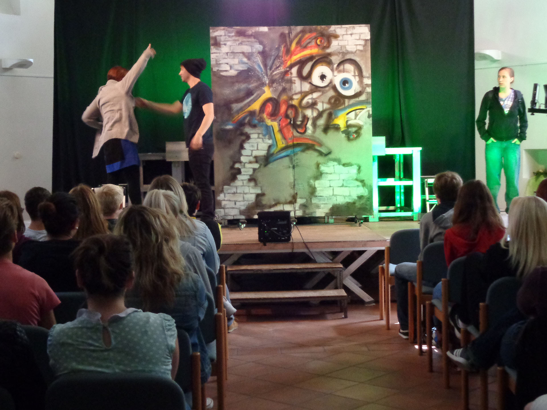 Theateraufführung im Rahmen des VIDETO-Projekts zur Gewaltprävention und Mobbingbekämpfung: Auf der Bühne mit Graffiti-Backdrop interagieren Performer, während das Publikum zuschaut.