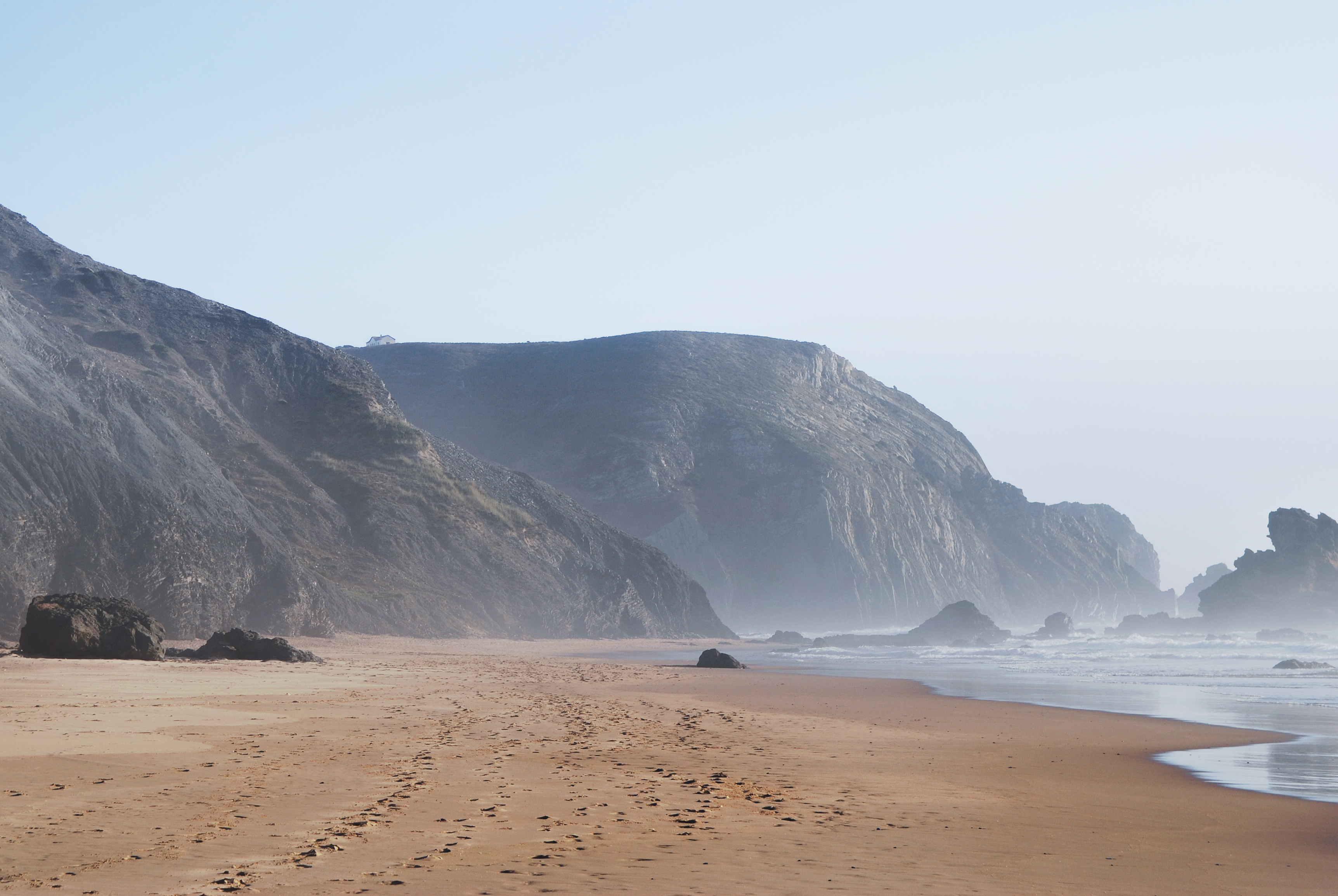 Ein leerer Sandstrand mit Fußspuren, umgeben von steilen felsigen Klippen und dem Meer, unter einem hellen Himmel.
