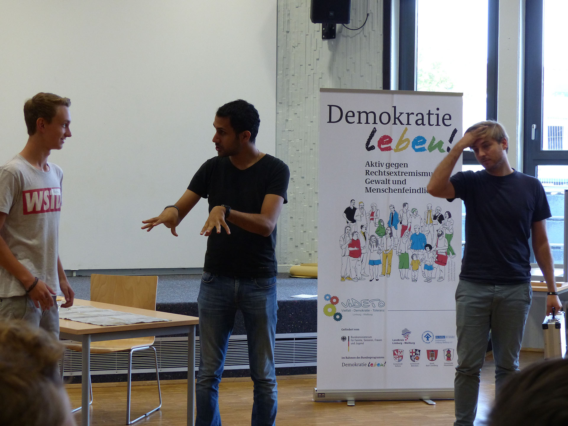 Drei Personen stehen in einem Raum mit einem Banner, das „Demokratie leben!“ und das VIDETO-Logo trägt, während sie an einer Aktivität zum interkulturellen Lernen und der Stärkung demokratischer Werte teilnehmen, die Willkommenskultur fördert.