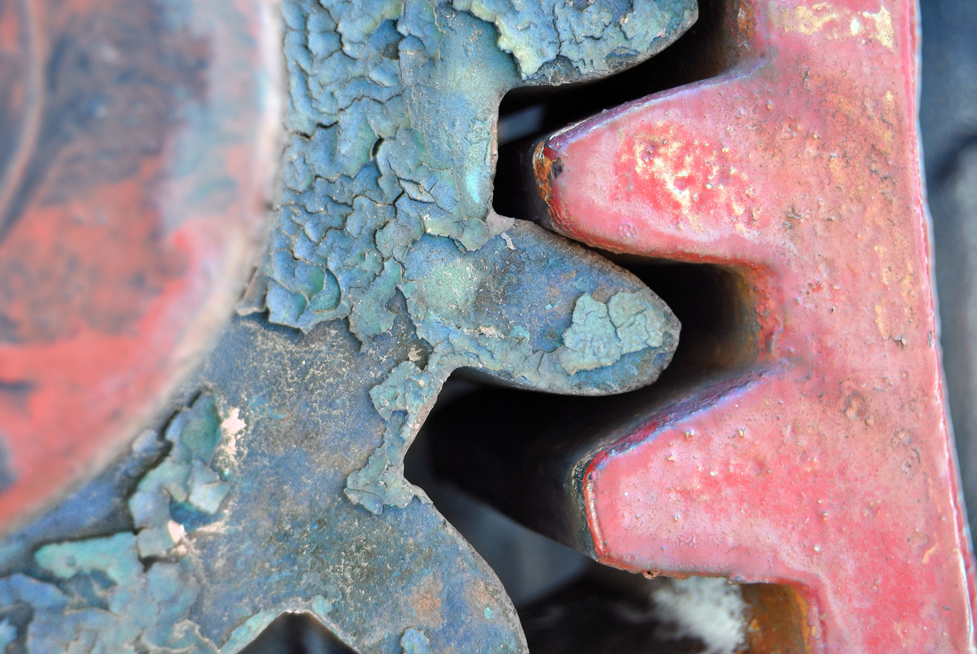 Nahaufnahme eines Zahnrads mit abblätternder blau-grüner Patina und roter Farbschicht, das mechanische Strukturen und Alterungserscheinungen zeigt.