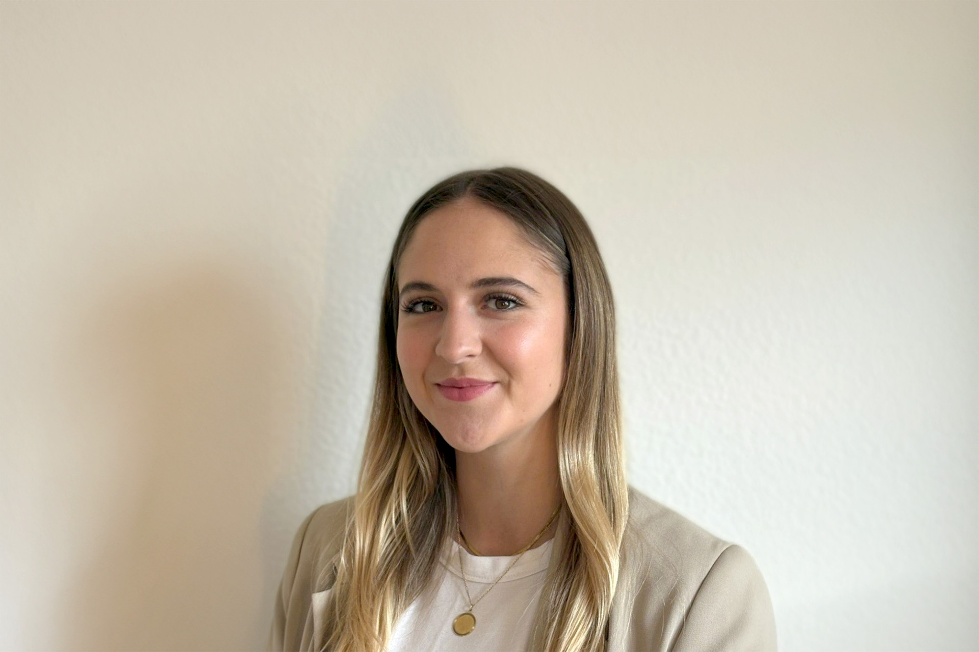 Chiara Kröller, Teil des VIDETO-Teams, mit langen blonden Haaren, beigem Blazer und weißem Top, steht vor einer hellen Wand.