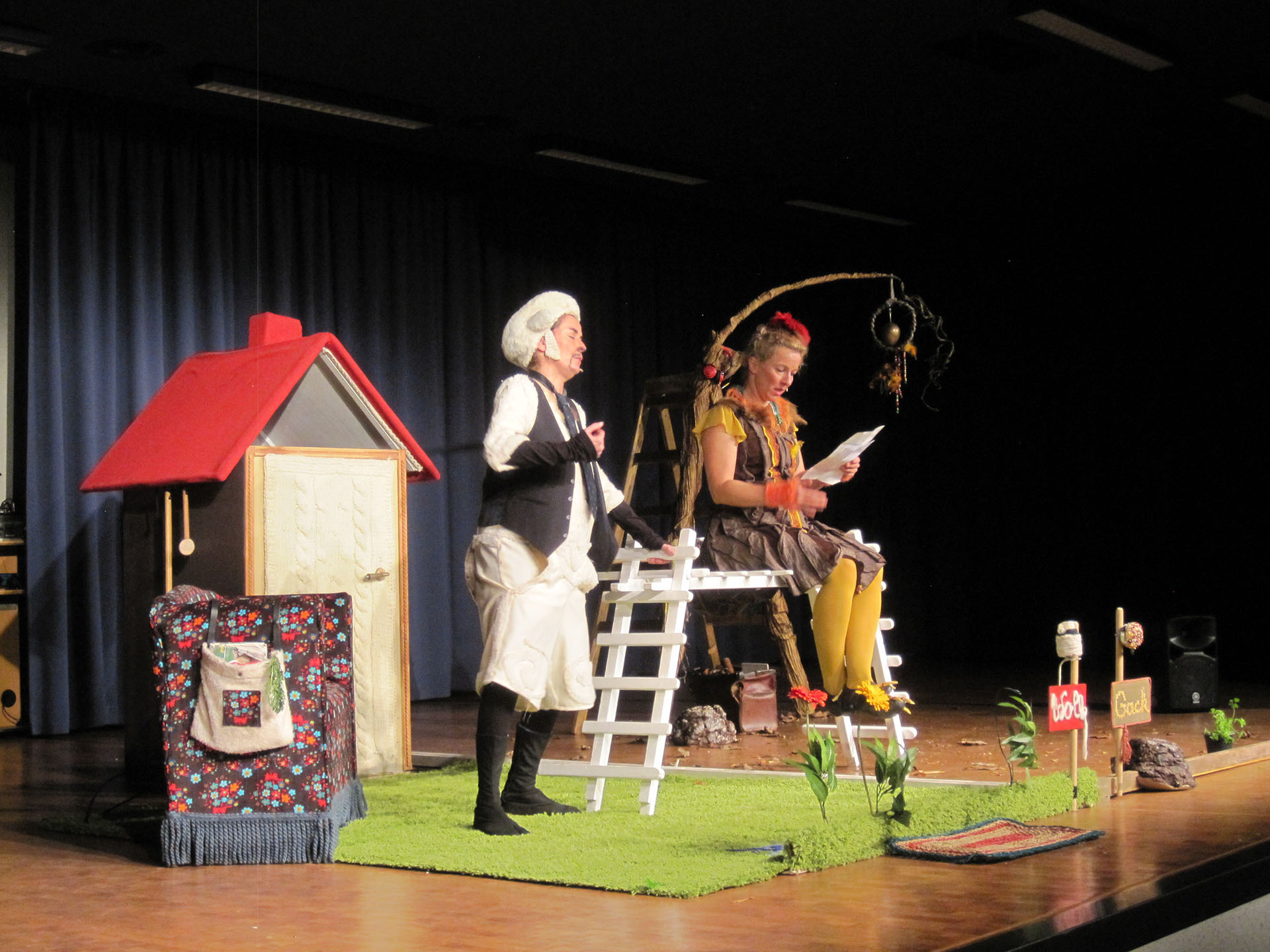Bühnenaufführung mit zwei Schauspielern in Kostümen, die vor einem kleinen Haus mit rotem Dach, einer Leiter und Pflanzenrequisiten agieren – Teil eines Projekts zur Förderung von Demokratie, Vielfalt und Toleranz.