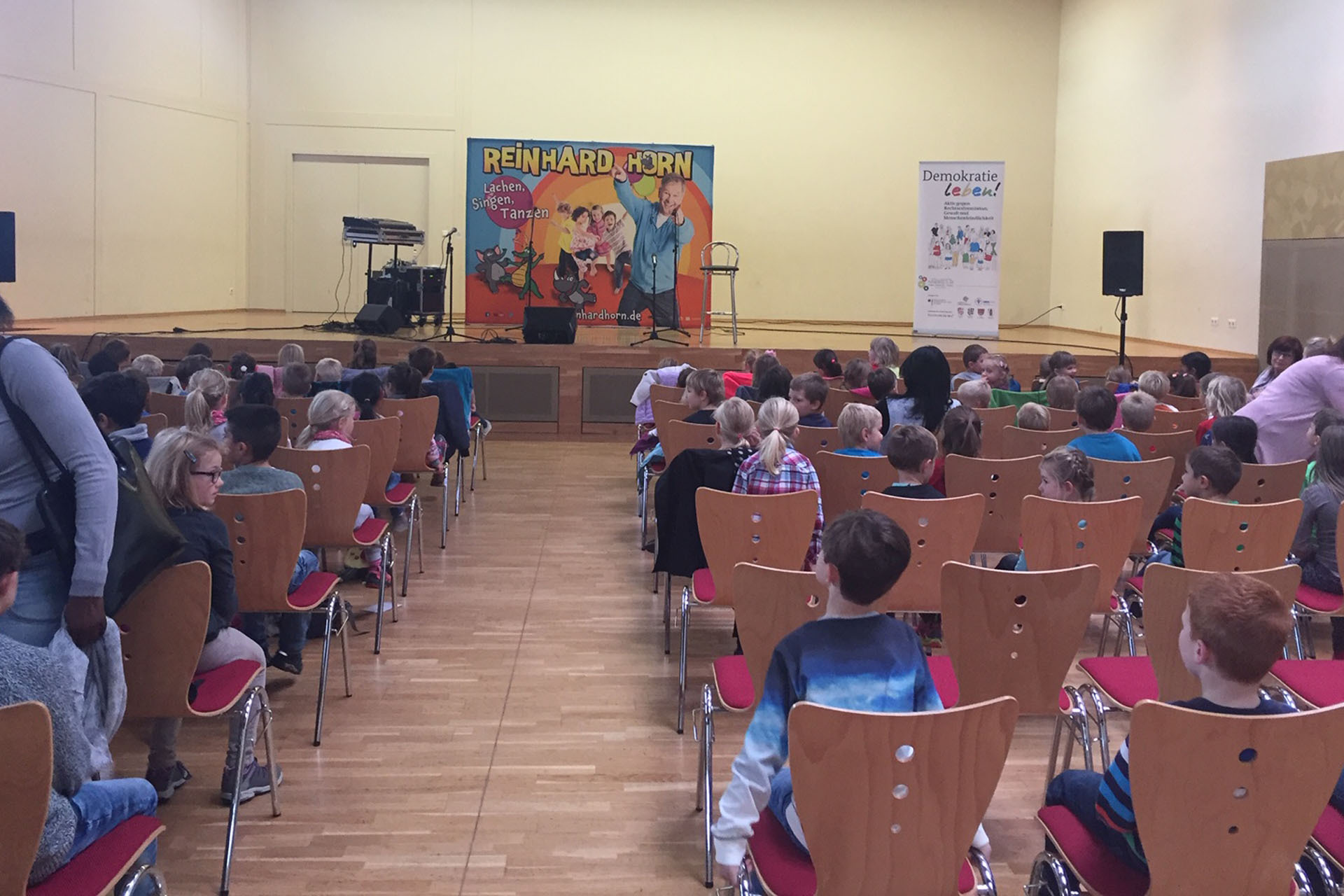 Ein Raum mit Kindern und Erwachsenen bei einem Bühnenauftritt von Reinhard Horn (Banner: 