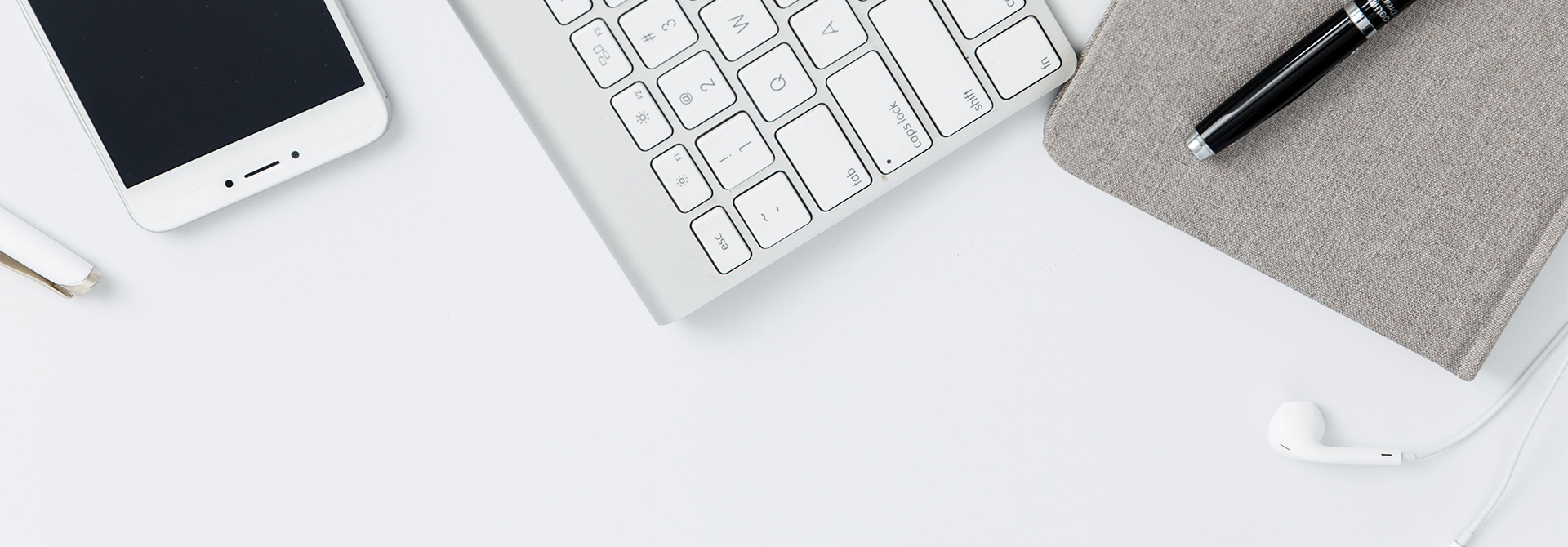Ein übersichtliches Arbeitsplatzsetup mit einer weißen Tastatur, einem Smartphone, einem grauen Notizbuch mit einem schwarzen Stift und weißen Kopfhörern auf einem hellen Untergrund.