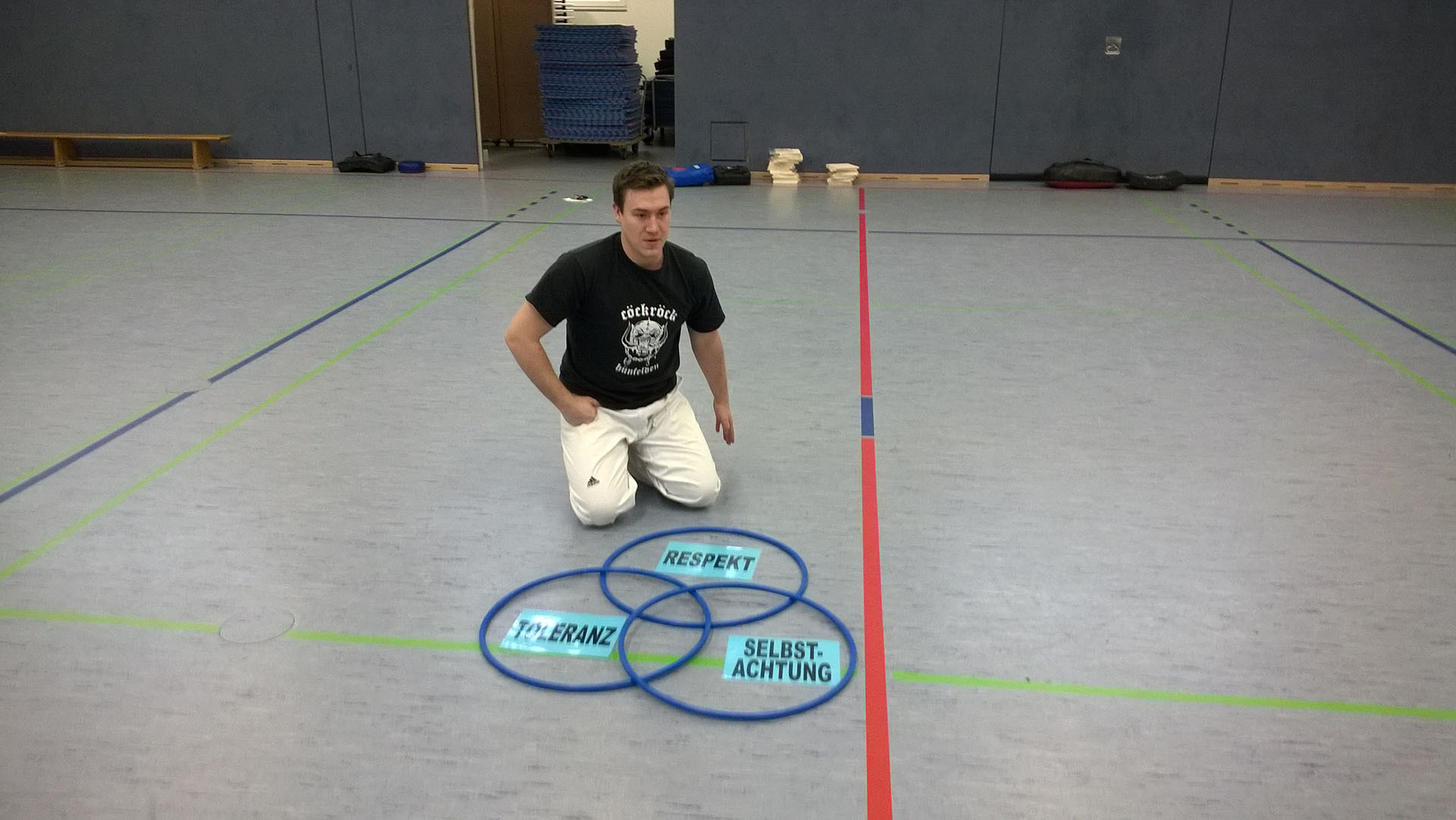 Ein Mann kniet in einer Sporthalle vor drei blauen Hula-Hoops, die mit den Begriffen „Toleranz“, „Respekt“ und „Selbstachtung“ beschriftet sind – ein Bild zur Förderung demokratischer Werte im Rahmen des VIDETO-Projekts.